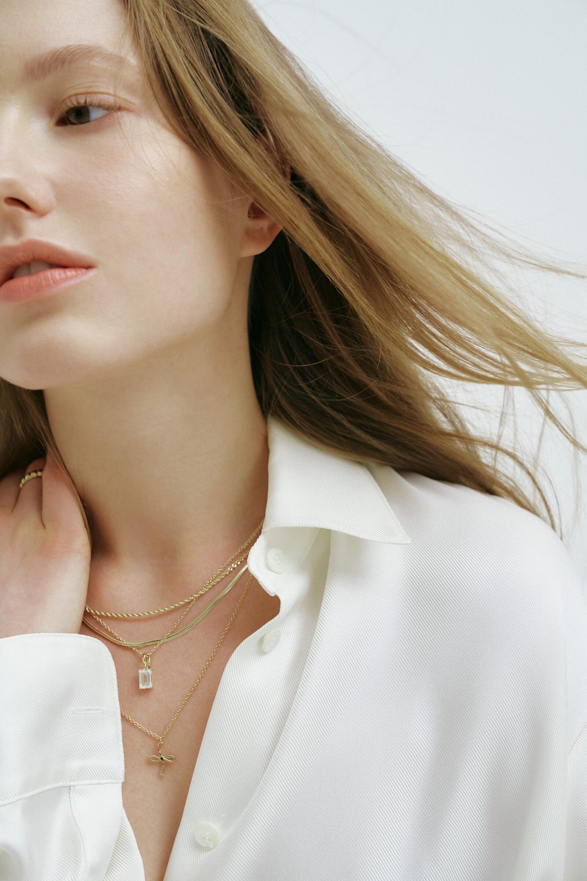A woman with her hair blowing to one side, hand at her neck, wearing a rope chain, a herringbone chain and two pendants.
