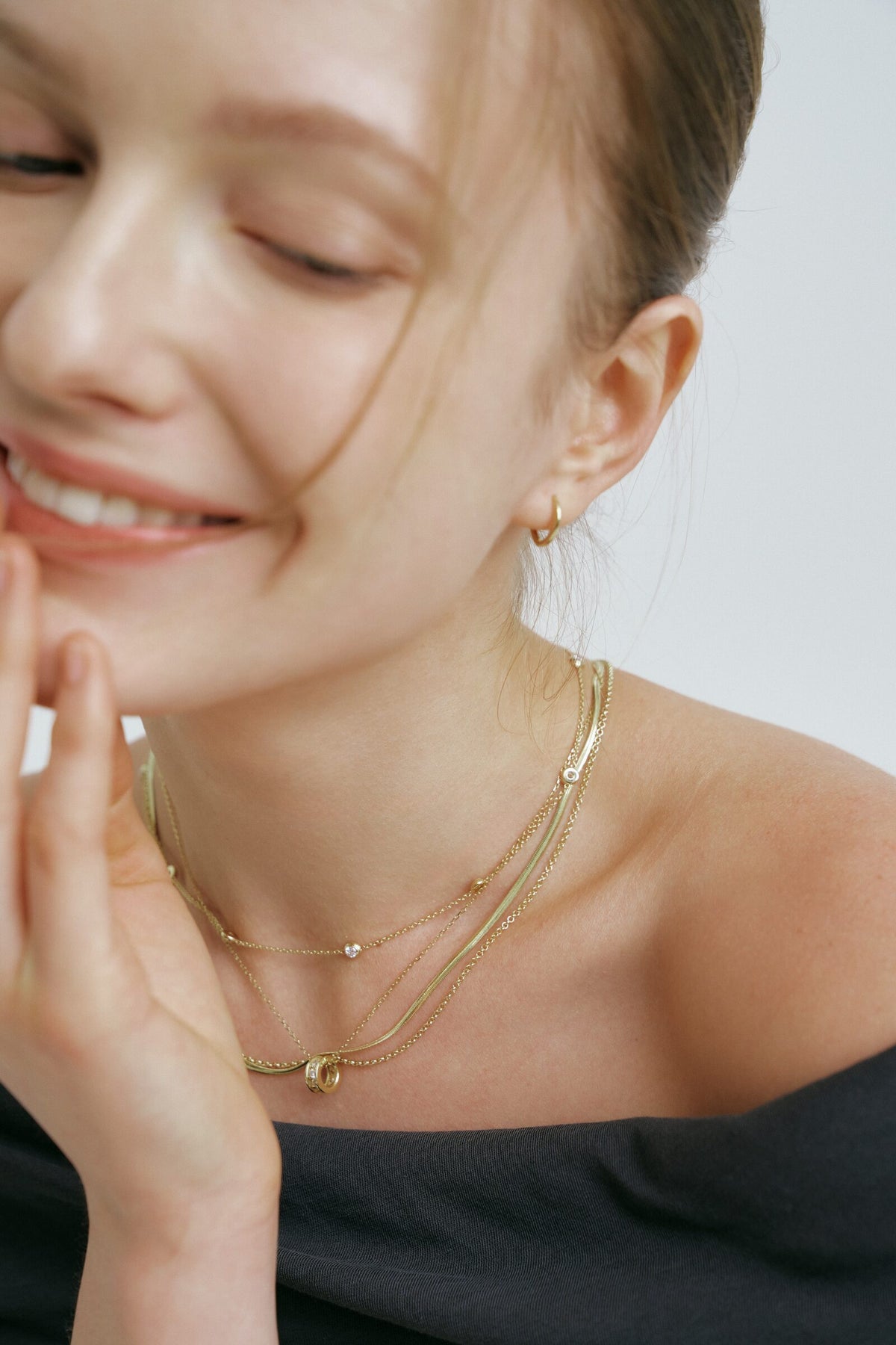 Smiling with a hand at her cheek, hair in a bun, thin gold hoops and layered chains over a charcoal off-shoulder top.