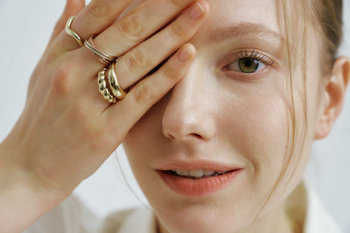 Close frame: one hand over her eye, gold dome rings stacked on her fingers.
