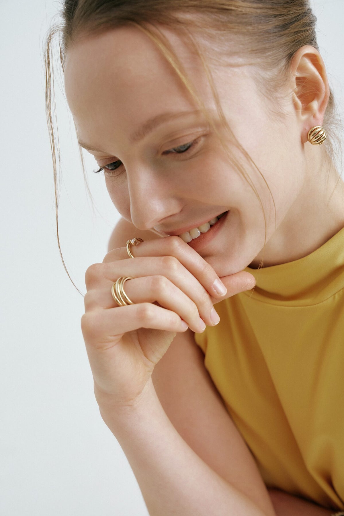 A woman smiles down with her hand at her chin, a coiled ring, a ring with a clear stone and a ridged dome earring.