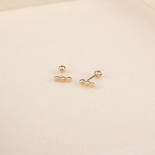 Dainty yellow gold bar stud earrings with three ball details and rounded screw-back posts on beige surface