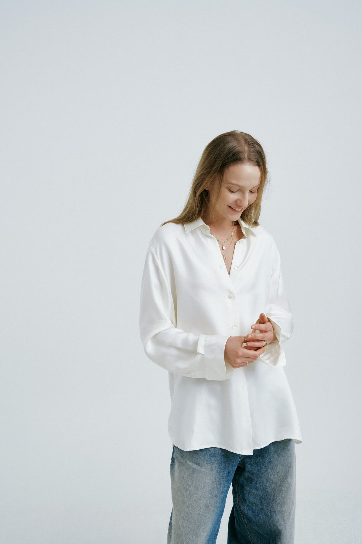 Full figure in an oversized white shirt and jeans, standing alone in the studio, layered gold necklaces.