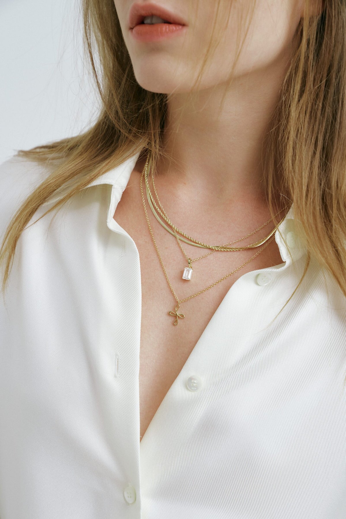 Close crop at the open collar: an emerald-cut stone pendant, the Bloom Necklace and a herringbone chain layered together.