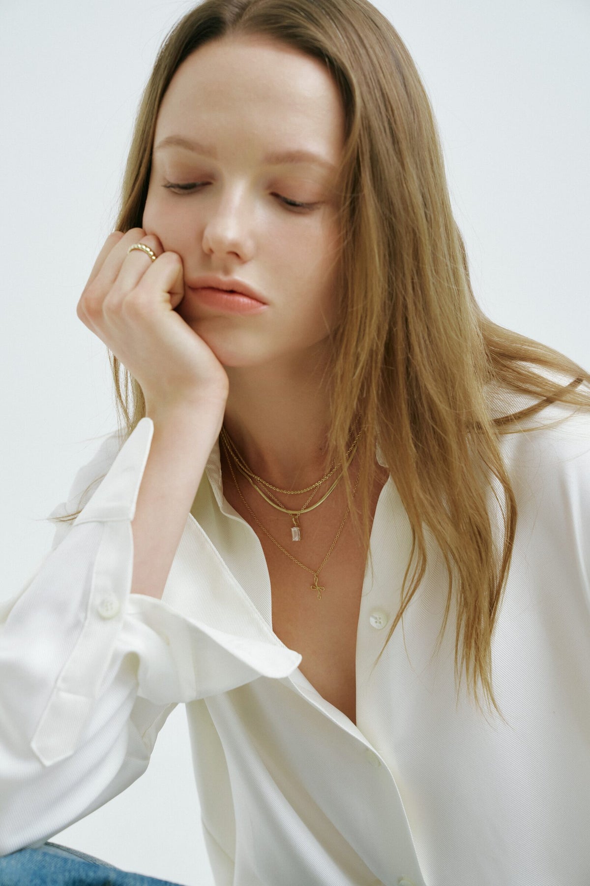 A woman rests her cheek on her hand, eyes closed, a ridged ring and chains with a clear stone and a cross pendant.