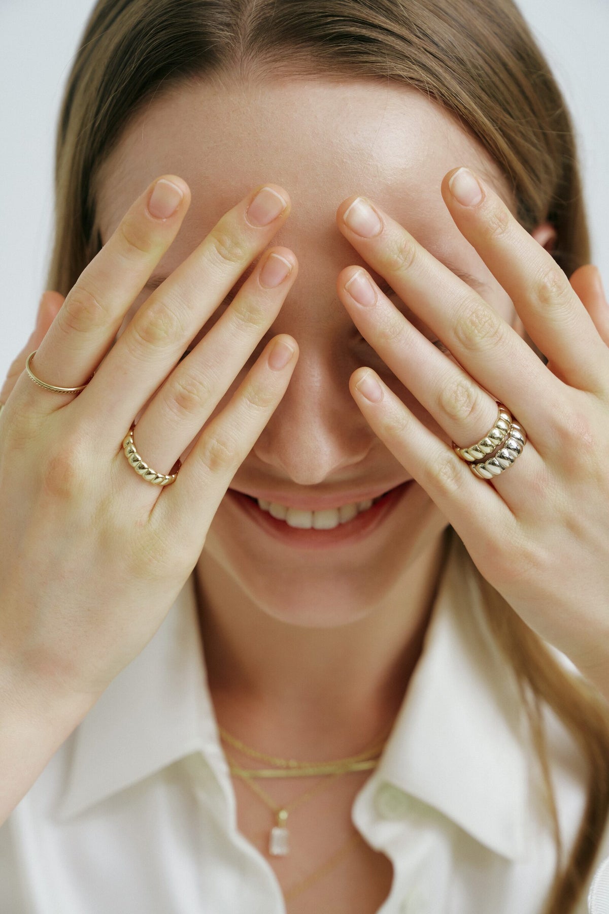 A woman covers her eyes with both hands and smiles, ridged rings on her fingers and layered chains at her collar.