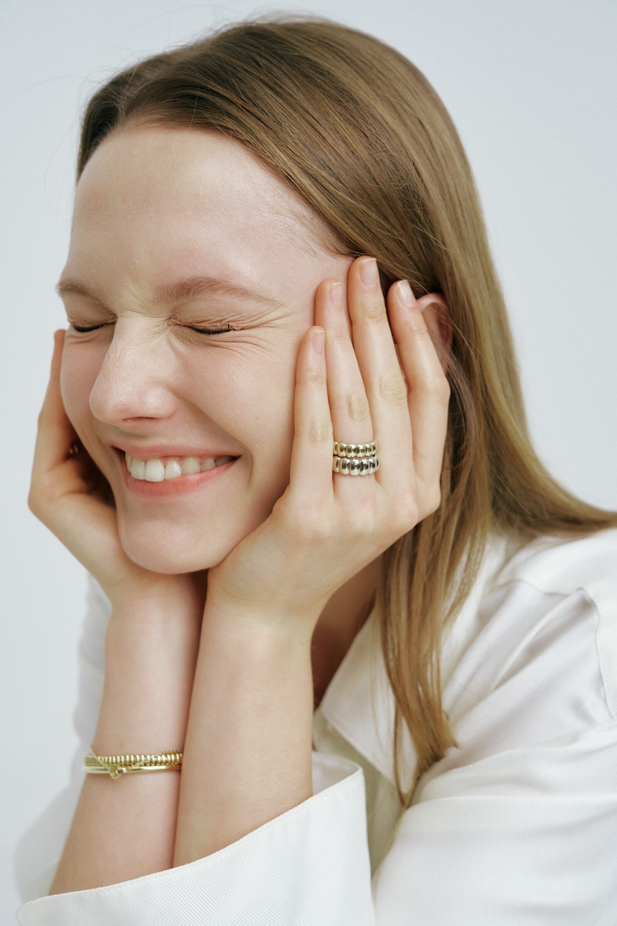 Eyes shut and laughing, hands to her cheeks, a Pillow Ring stack and the Accordion Bracelet.