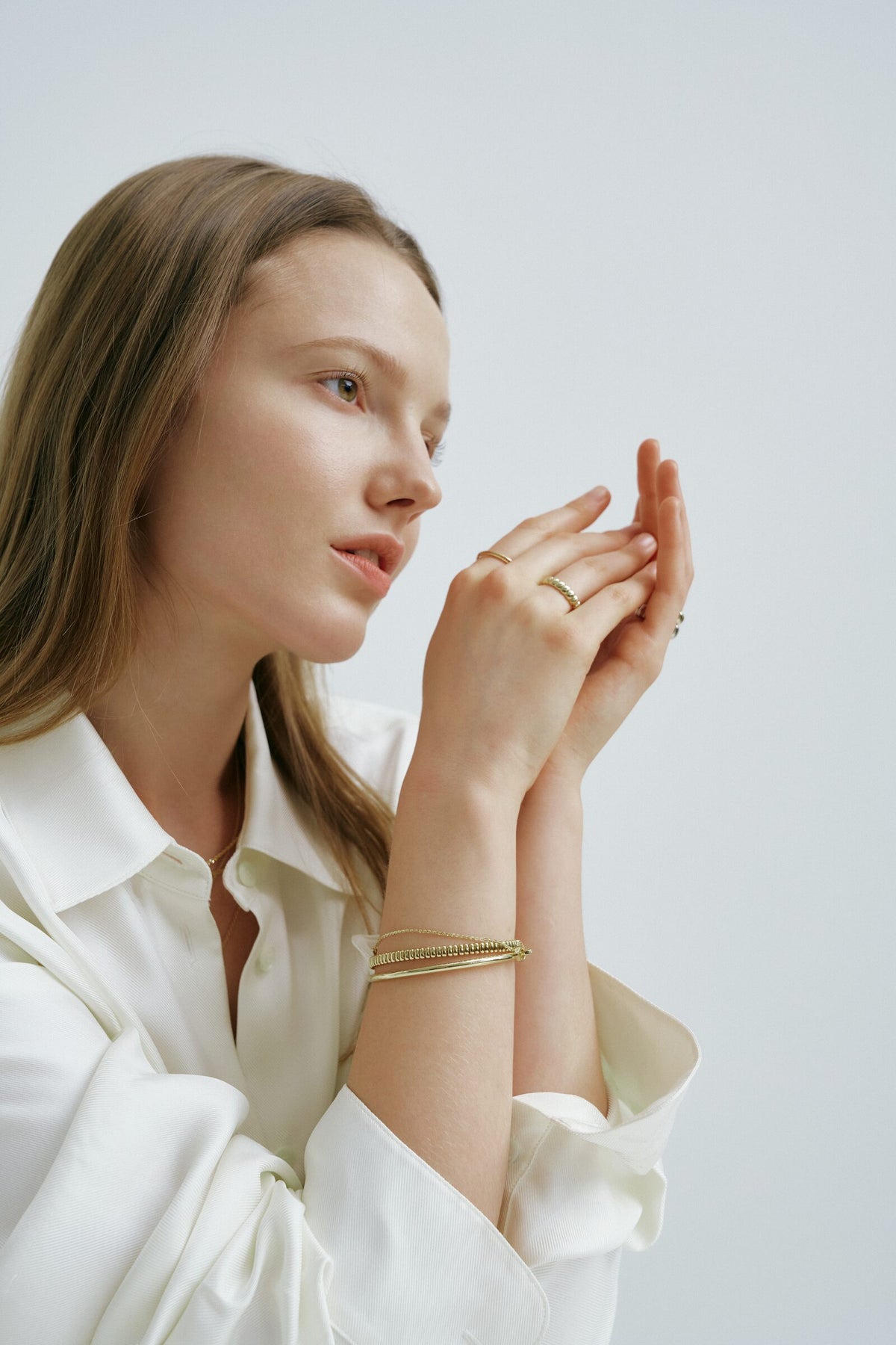 A woman in profile raises her hands toward her face, slim rings on her fingers, an Accordion Bracelet, a Classic Bangle and a fine chain bracelet at her wrist.