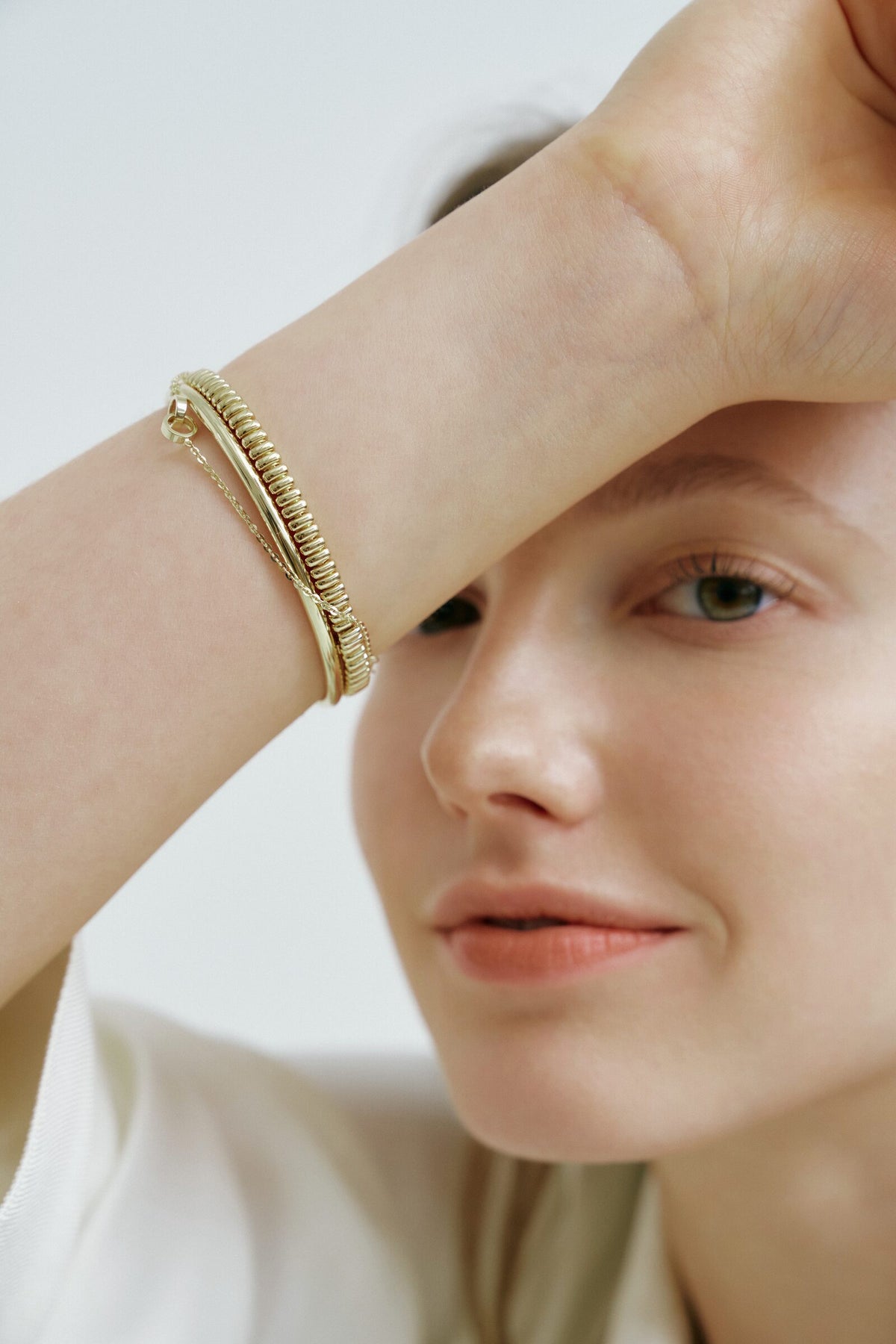 A woman rests her forearm on her forehead, a ridged bangle, a smooth bangle and a chain bracelet stacked at her wrist.