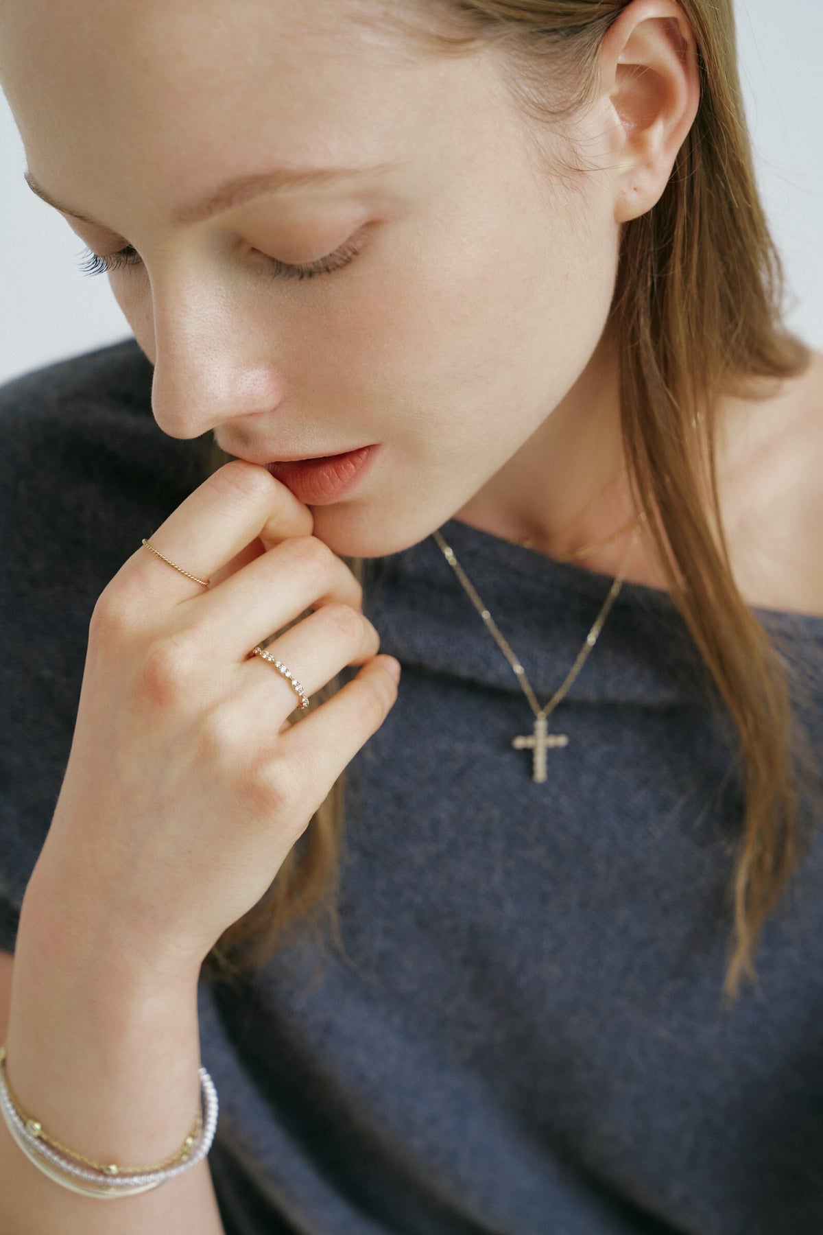A woman looks down with her fingers at her lips, a cross pendant on a chain, two slim rings and a bracelet of clear stones.