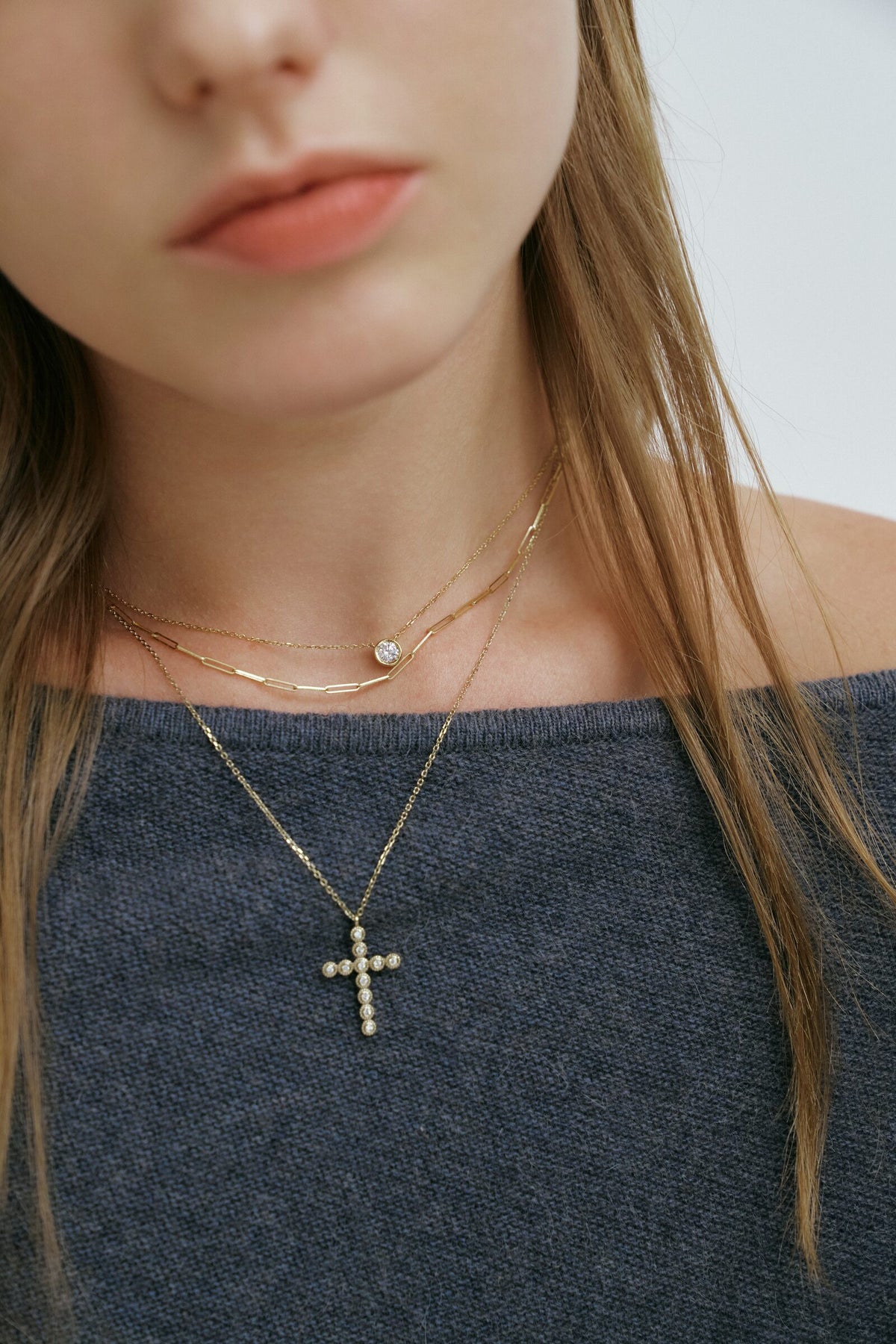 Close crop below the lips: the Bezel Cross Necklace over a slate knit, with a bezel solitaire and a link chain above it.