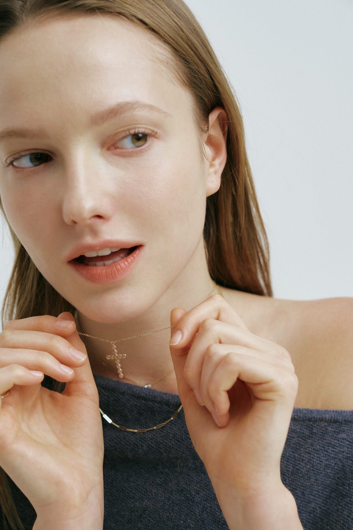 She adjusts a fine chain at her neck with both hands, mid-word, in a slate knit.