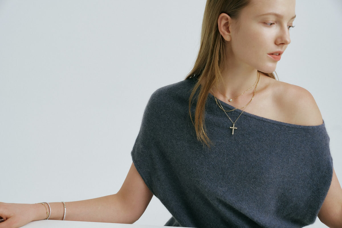 A woman in an off-the-shoulder knit looks to one side, a cross pendant and a short chain at her neck, bangles at her wrist.