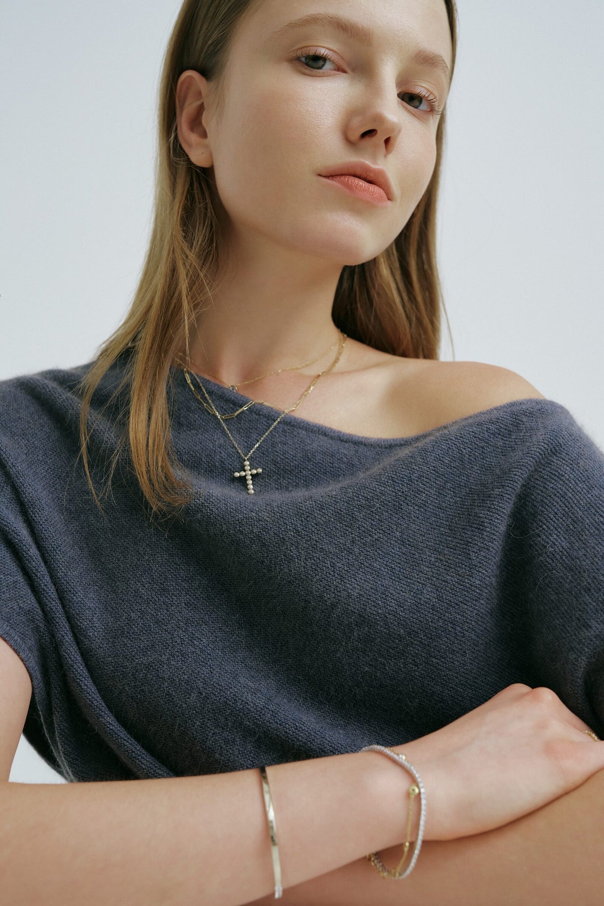 A woman with arms crossed looks into the camera, a cross pendant of clear stones, a bangle and a bracelet of clear stones.