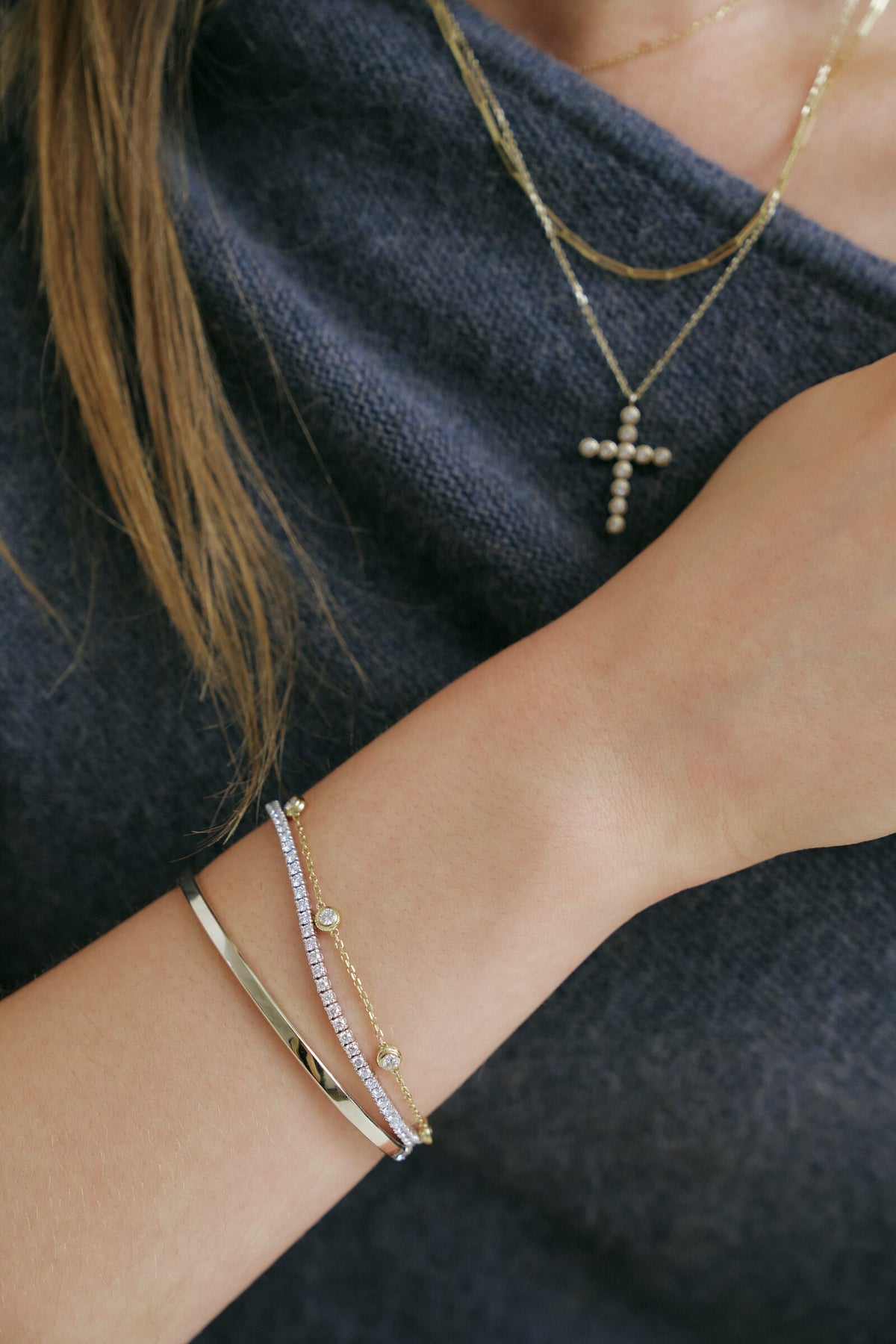 A forearm across the knit, a lab grown diamond tennis bracelet and a bezel station bracelet at the wrist, the cross pendant above.