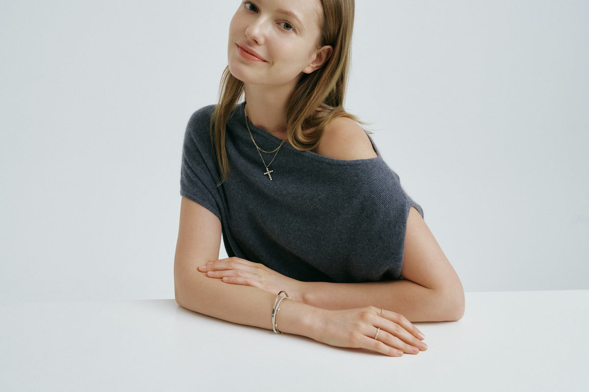 A woman leans on a table, arms folded, and smiles at the camera, a cross pendant at her neck, a bangle and slim rings.