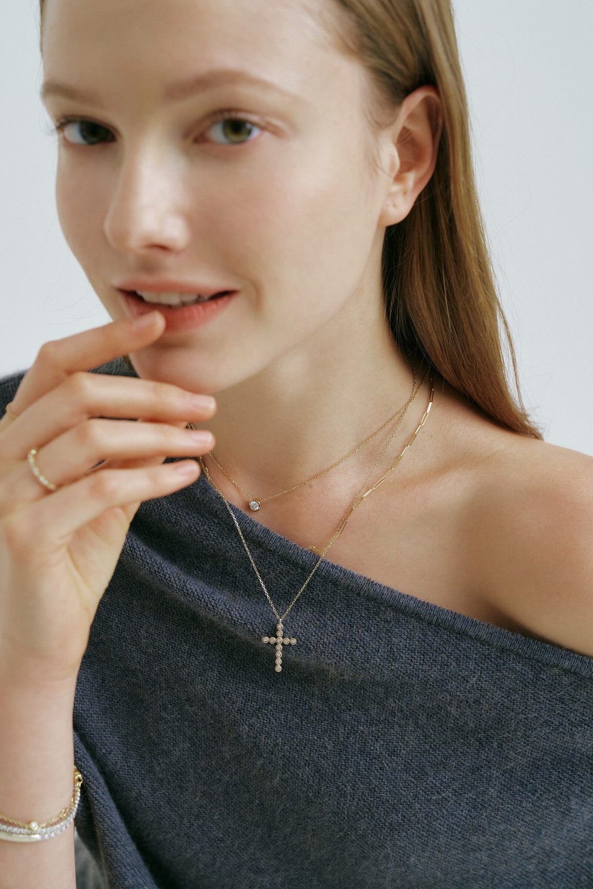A woman touches her lip and looks at the camera, a cross pendant of clear stones and a chain with one clear stone.