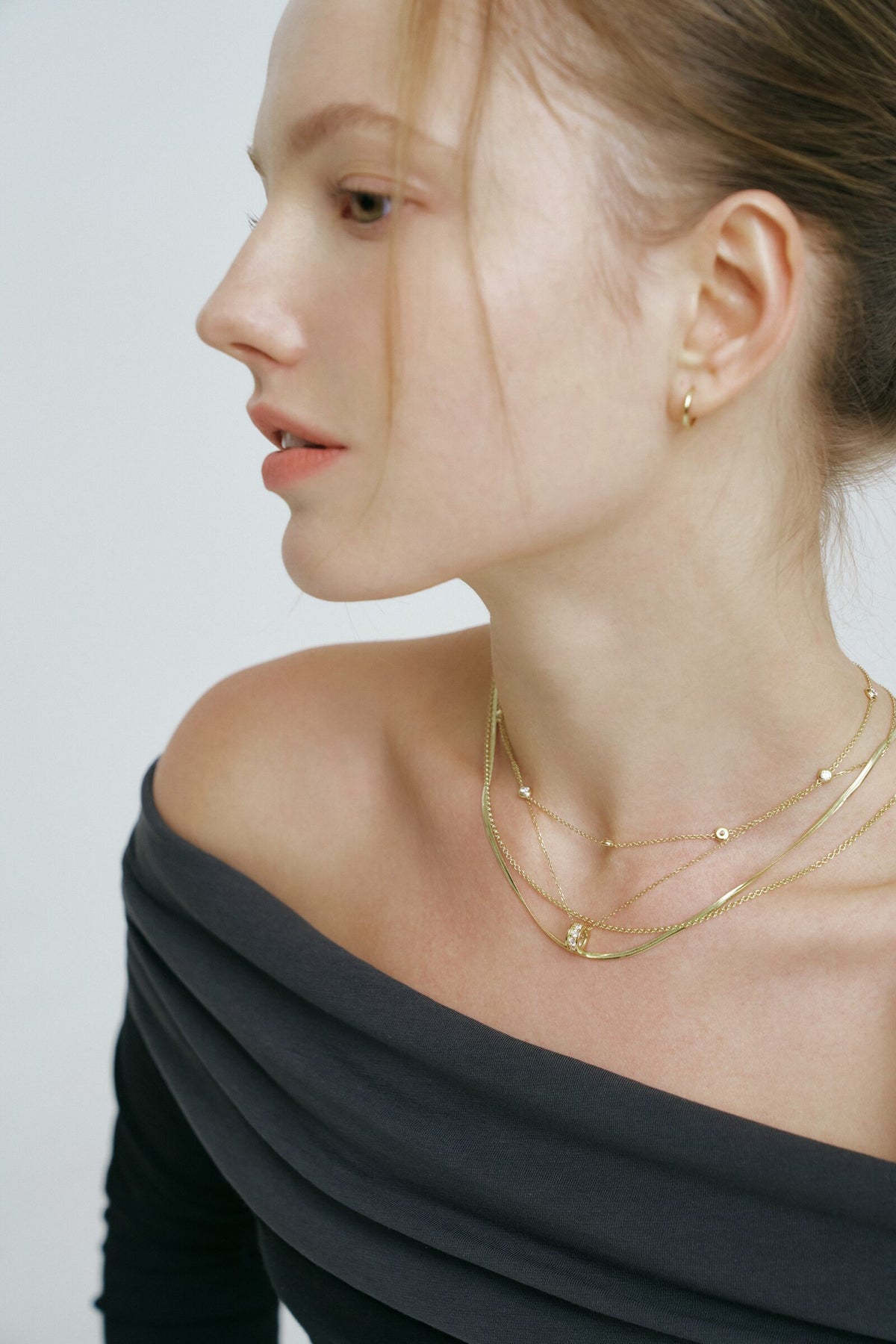In profile, hair in a bun, the Rondelle Necklace and layered chains above a charcoal neckline.