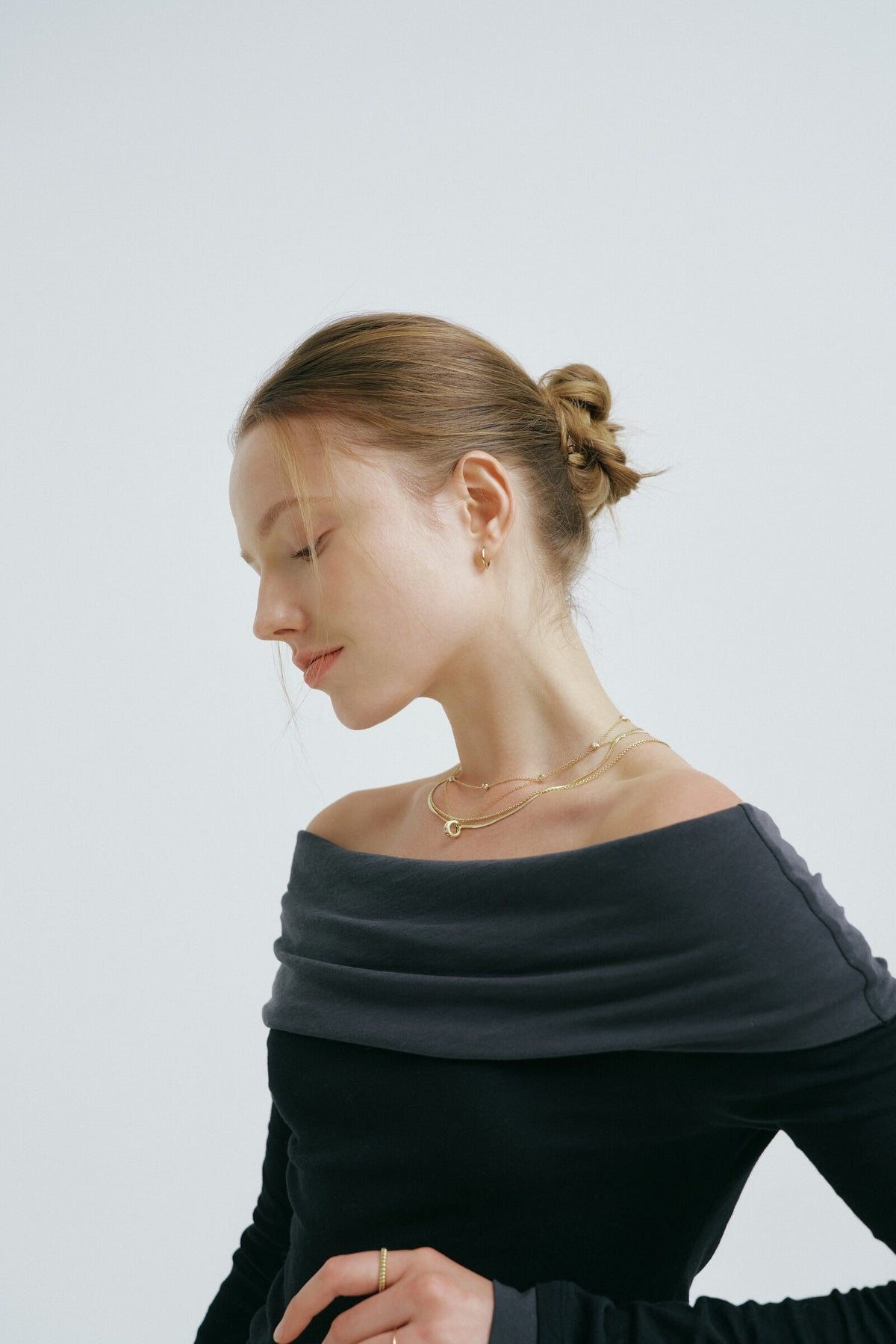 A woman in profile with her hair in a bun looks down, a hoop earring and layered chains with a round pendant at her neck.