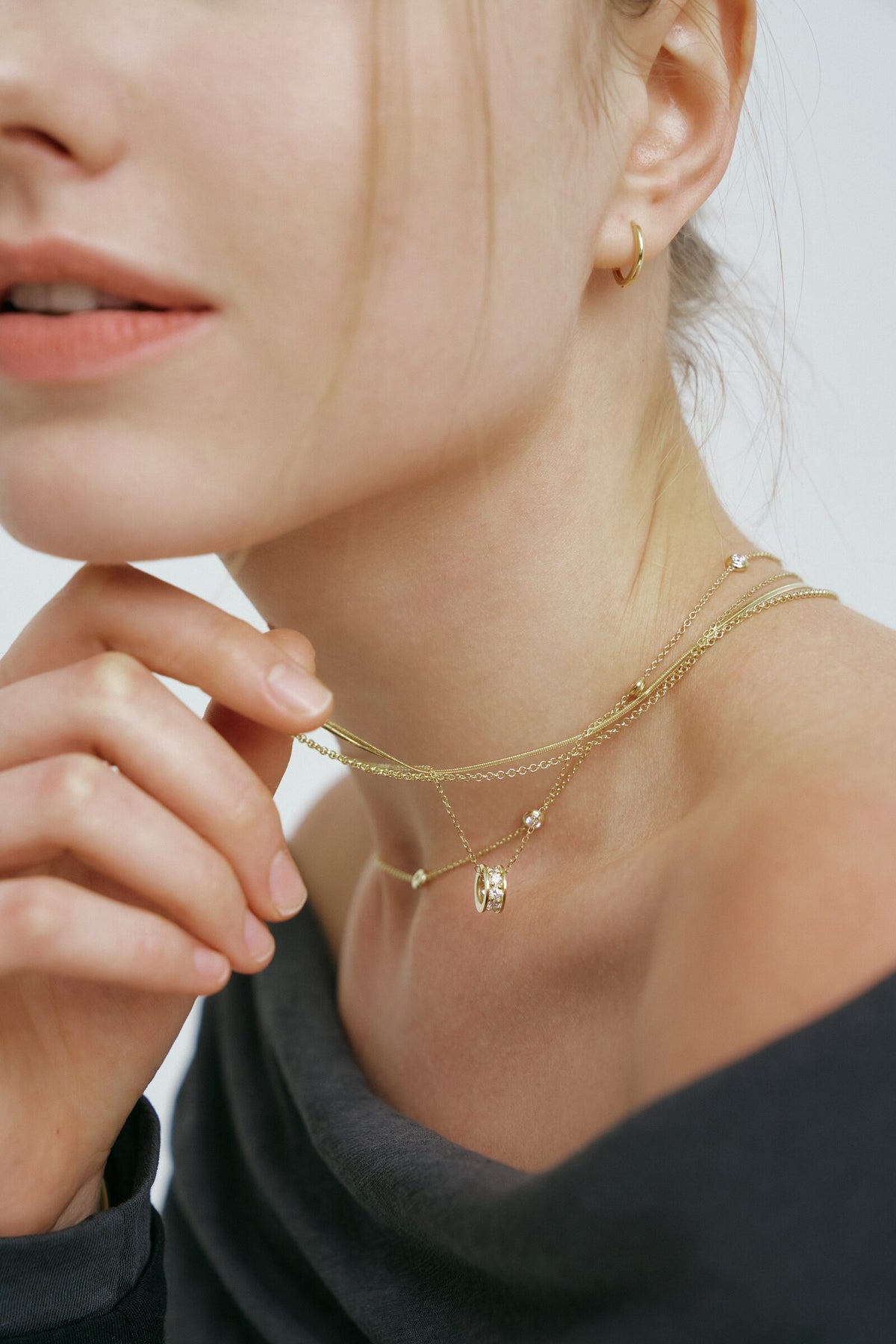 A woman touches her chin, seen close at the neck: a hoop earring and layered chains with clear stones and a ring pendant.