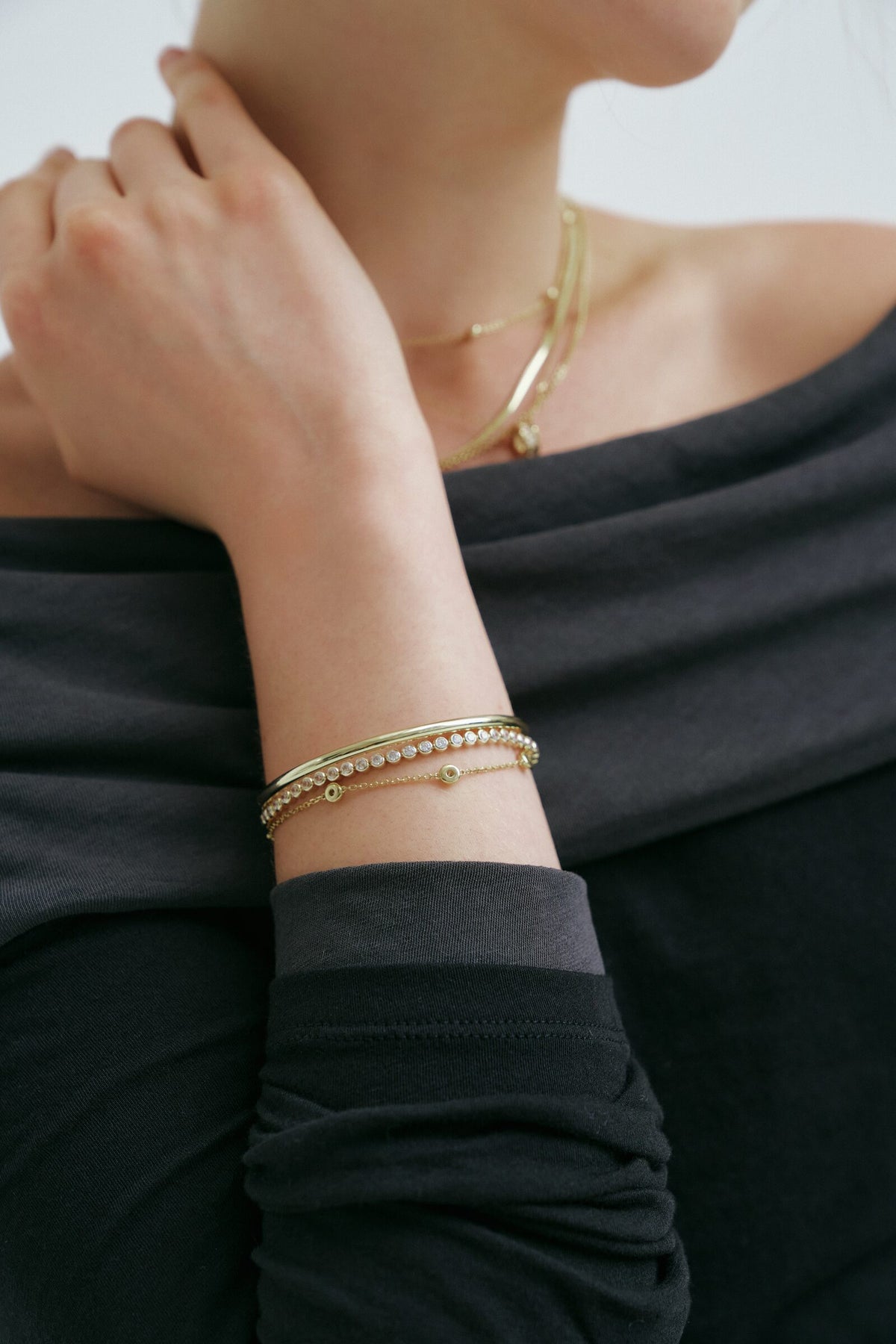A hand at the neck, the wrist stacked with a bangle, a bezel tennis bracelet and a bezel station chain.