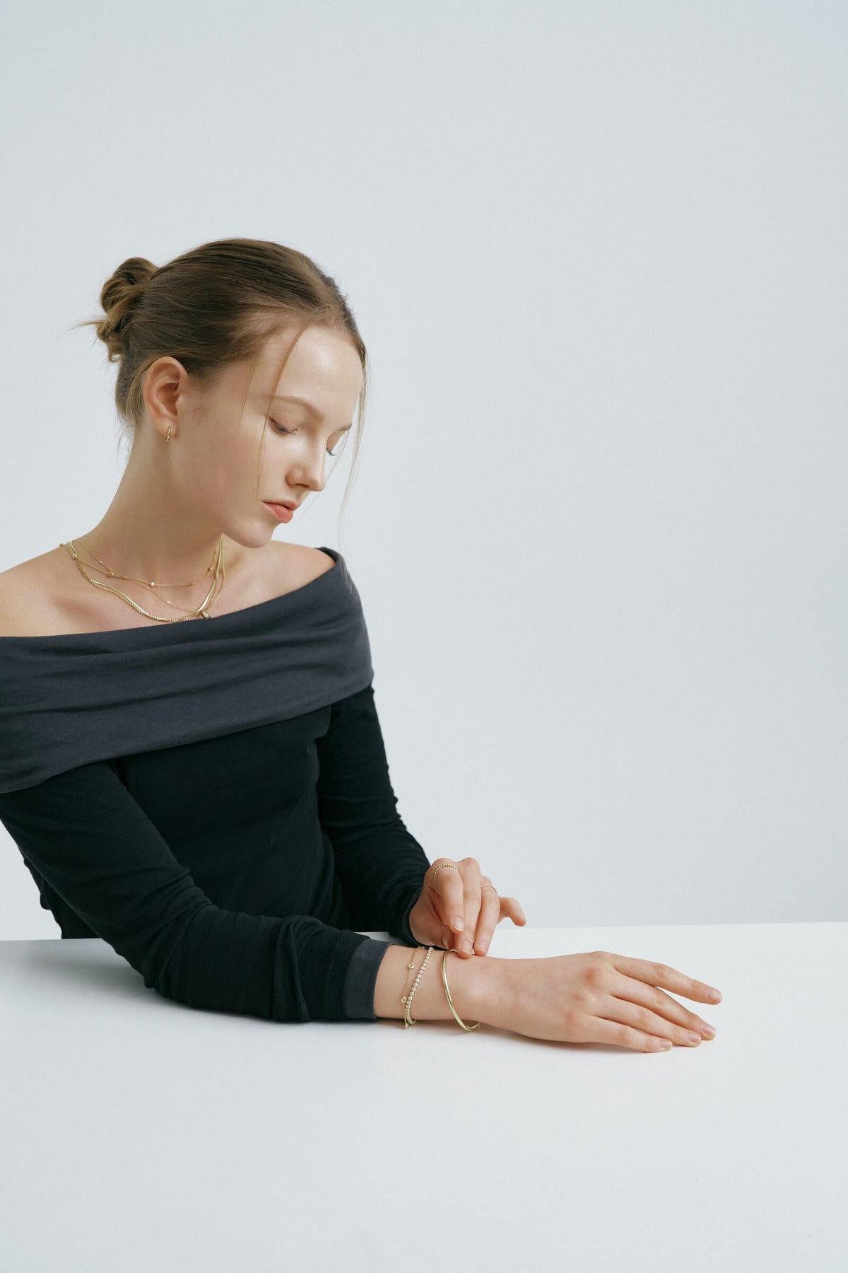 A woman seated at a table with her hair in a bun touches her wrist, layered chains at her neck and two bangles.