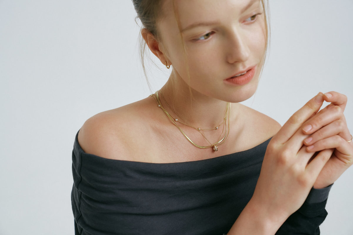 Wide frame: she glances down, one hand raised, layered necklaces over the charcoal top.