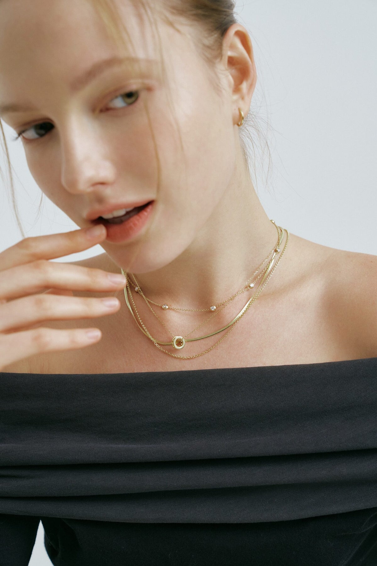 A woman with her hair pulled back touches her lip, a hoop earring and layered chains with clear stones and a round pendant.