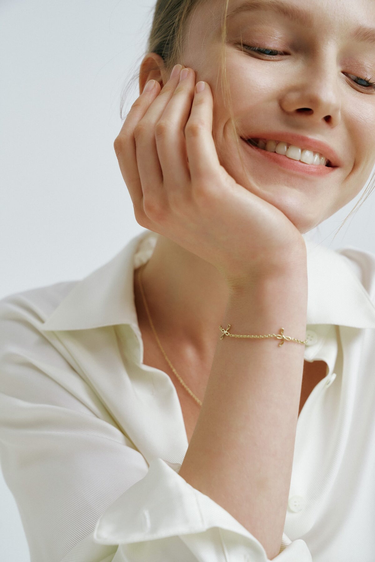 A woman laughs with her cheek in her hand, a chain bracelet with two small cross charms at her wrist.