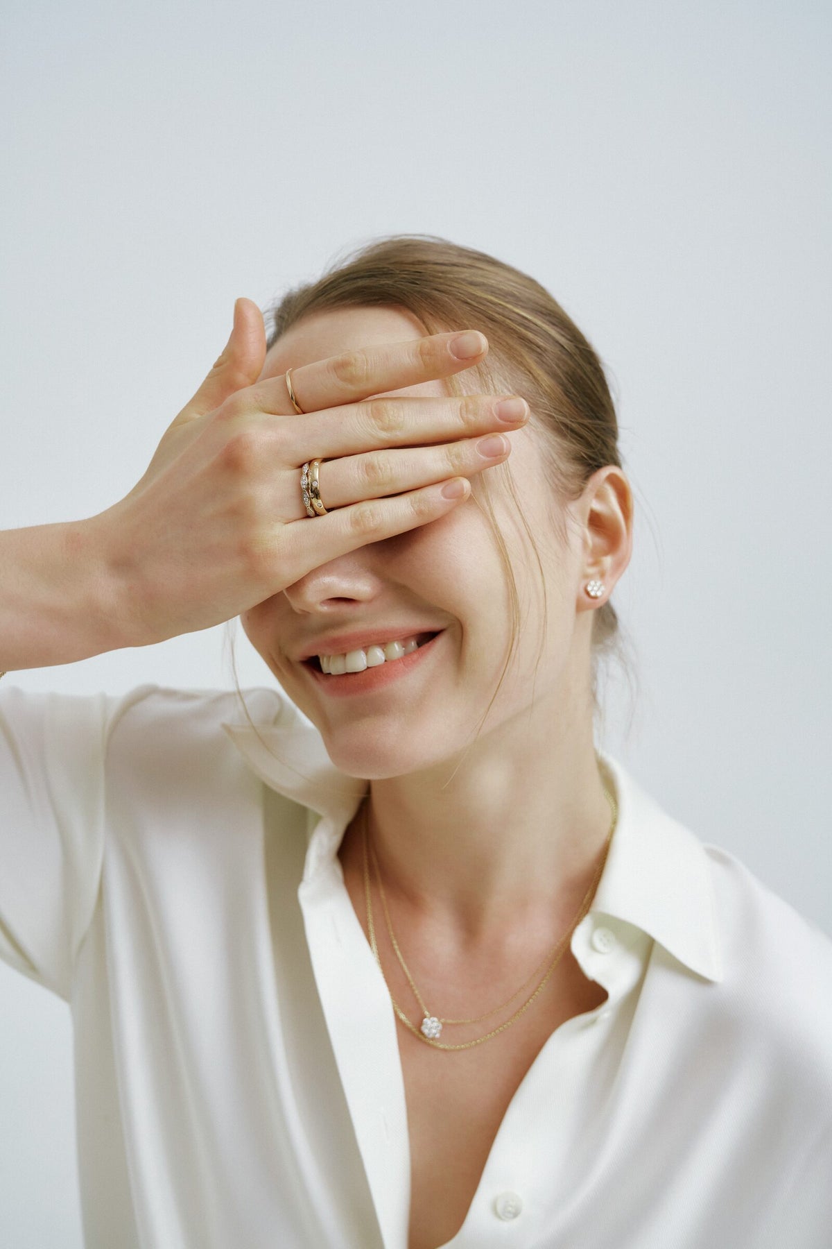 A woman covers her eyes with one hand and laughs, stacked rings, and a flower of clear stones at her ear and on a chain.