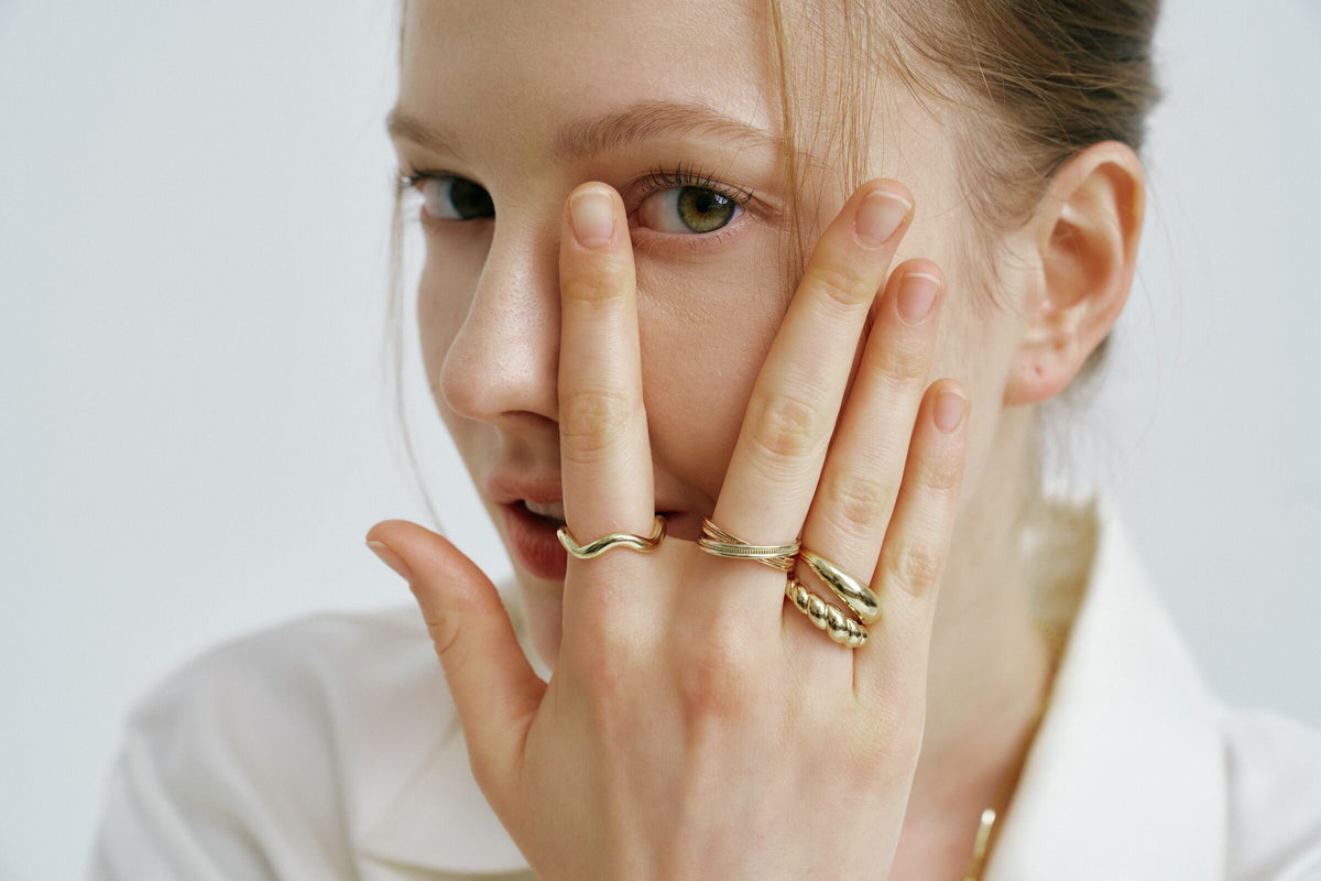 A woman covers half her face with one hand, a wavy ring, a braided ring, a domed ring and a ridged ring on her fingers.