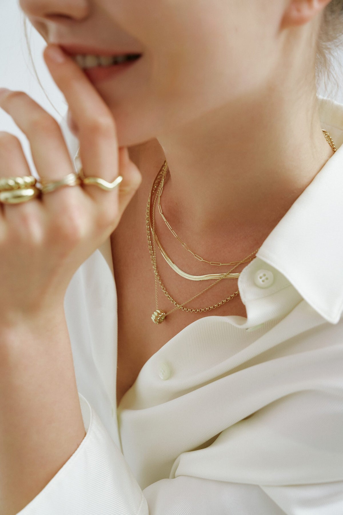 A woman smiles with a hand at her chin, ridged and wavy rings, a paperclip chain, a flat chain and a box chain.
