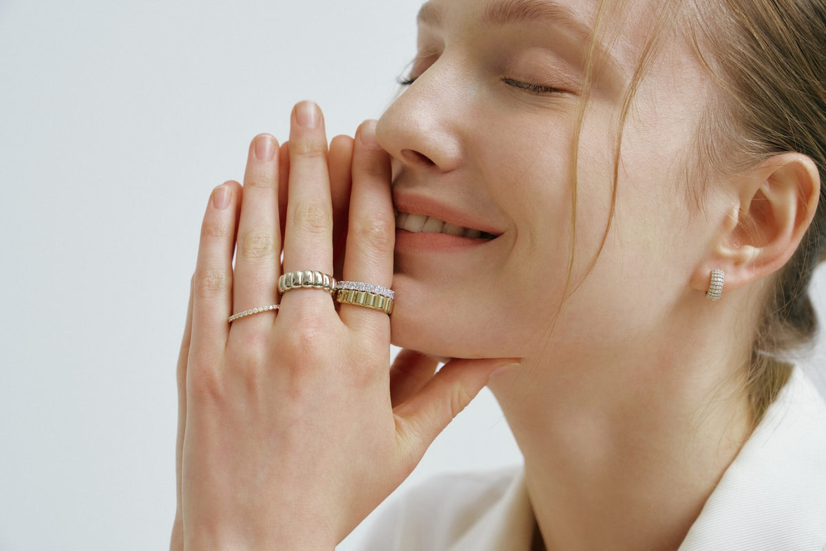 A woman smiles, eyes closed, hands pressed to her face, ridged rings, bands of clear stones and a hoop of clear stones.