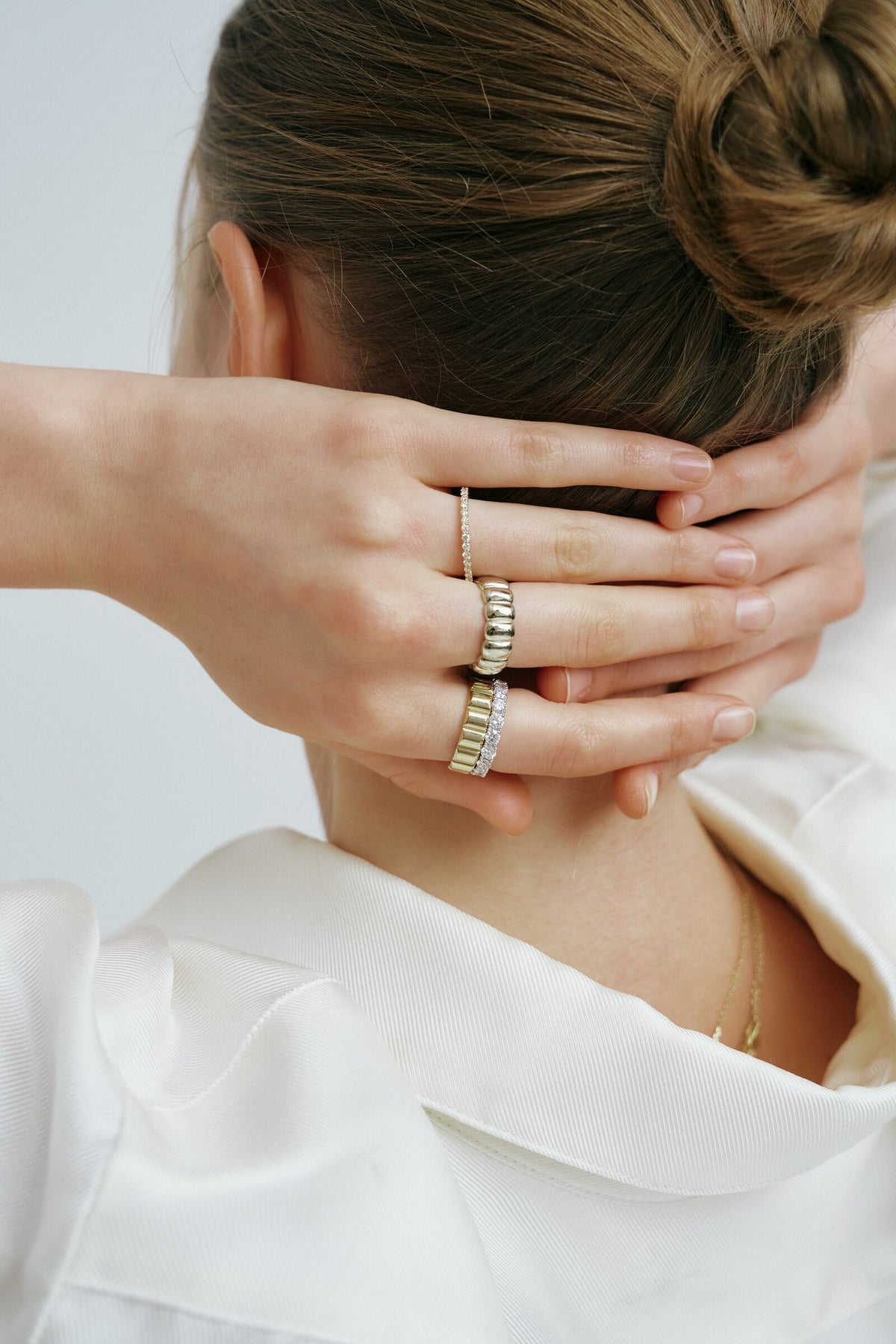 From behind, both hands at the nape below a bun, gold and diamond rings stacked.