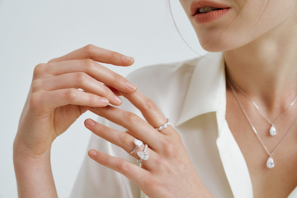 A woman lays one hand over the other, pear-shaped clear stones on two rings and on two pendant chains at her open collar.