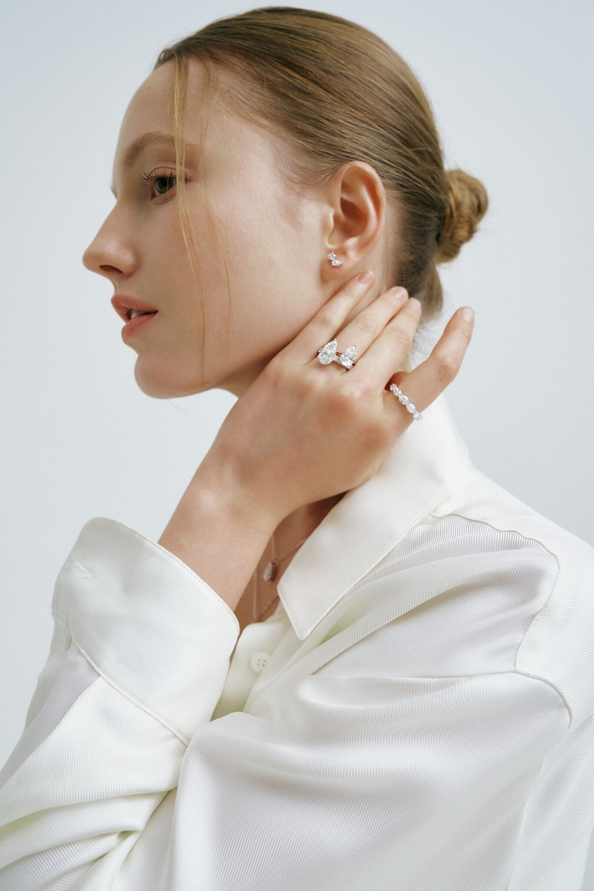 A woman in profile, hair in a bun, holds a hand at her neck, rings of oval and pear-shaped clear stones and a stud earring.