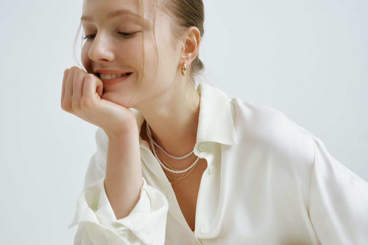 A woman smiles with her chin on her fist, a pearl strand and a line of clear stones layered with a chain at her open collar and a hoop earring.