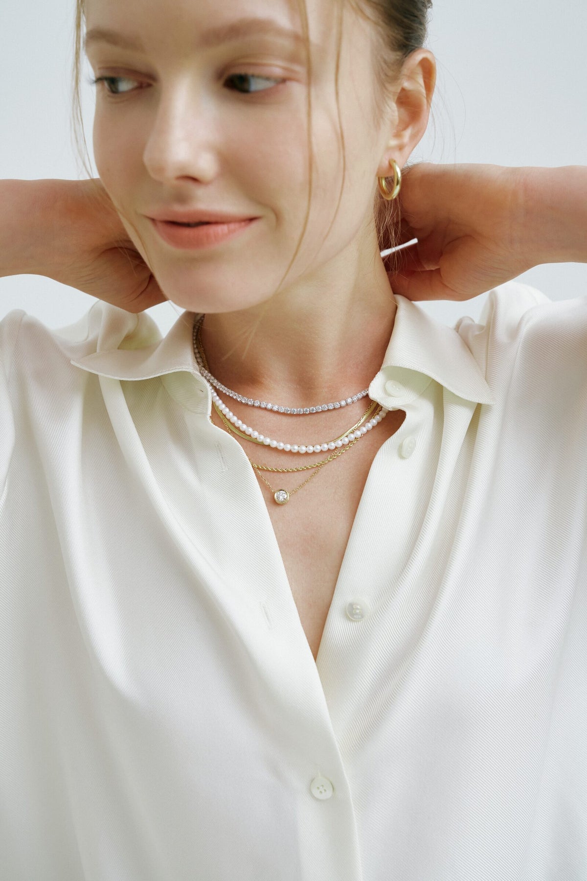 A woman lifts her hands behind her neck, a line of clear stones, a pearl strand and a chain with a round pendant.