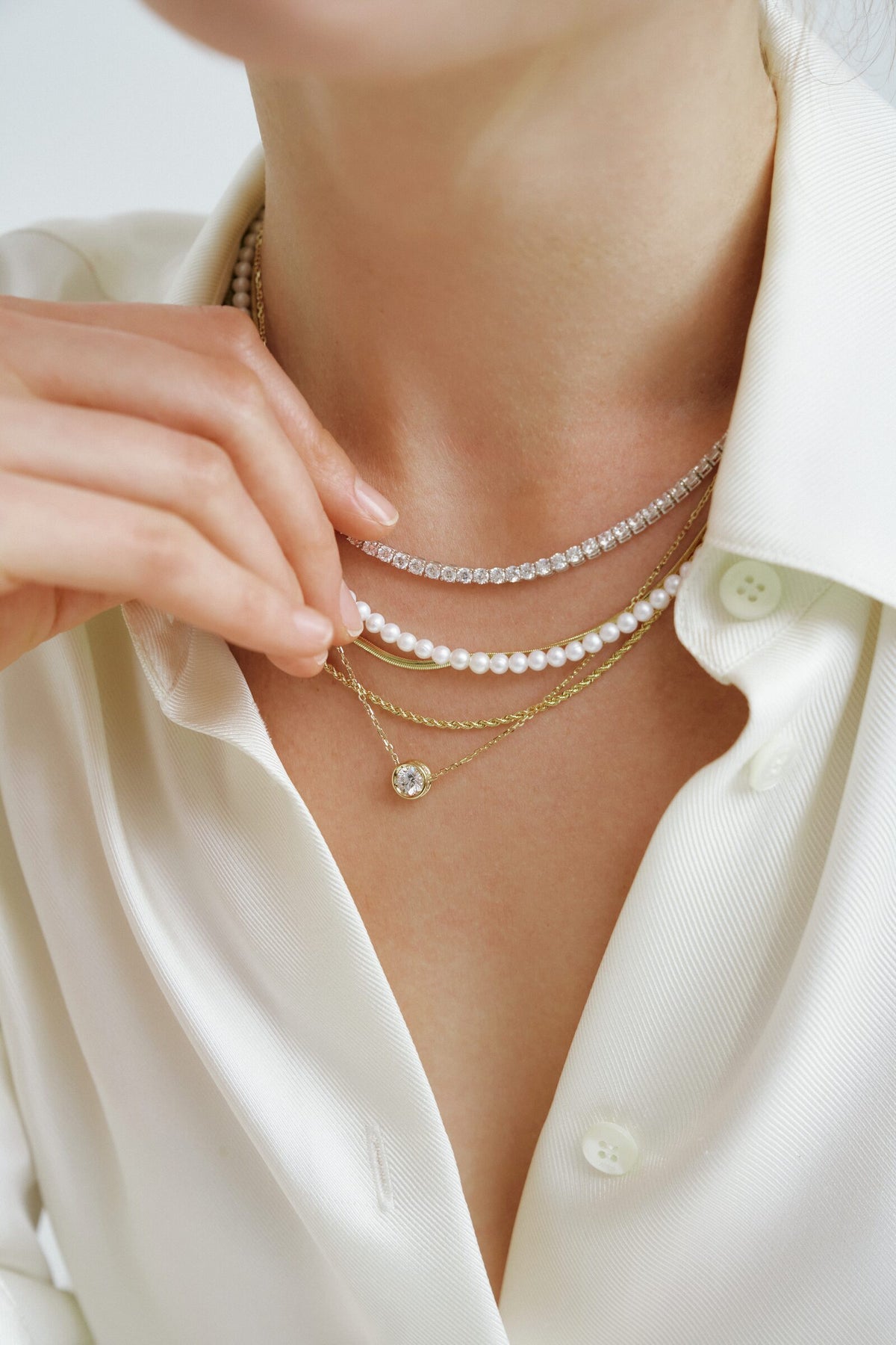 Fingers at the collar of a white shirt, a freshwater pearl strand layered with a diamond line and a solitaire pendant