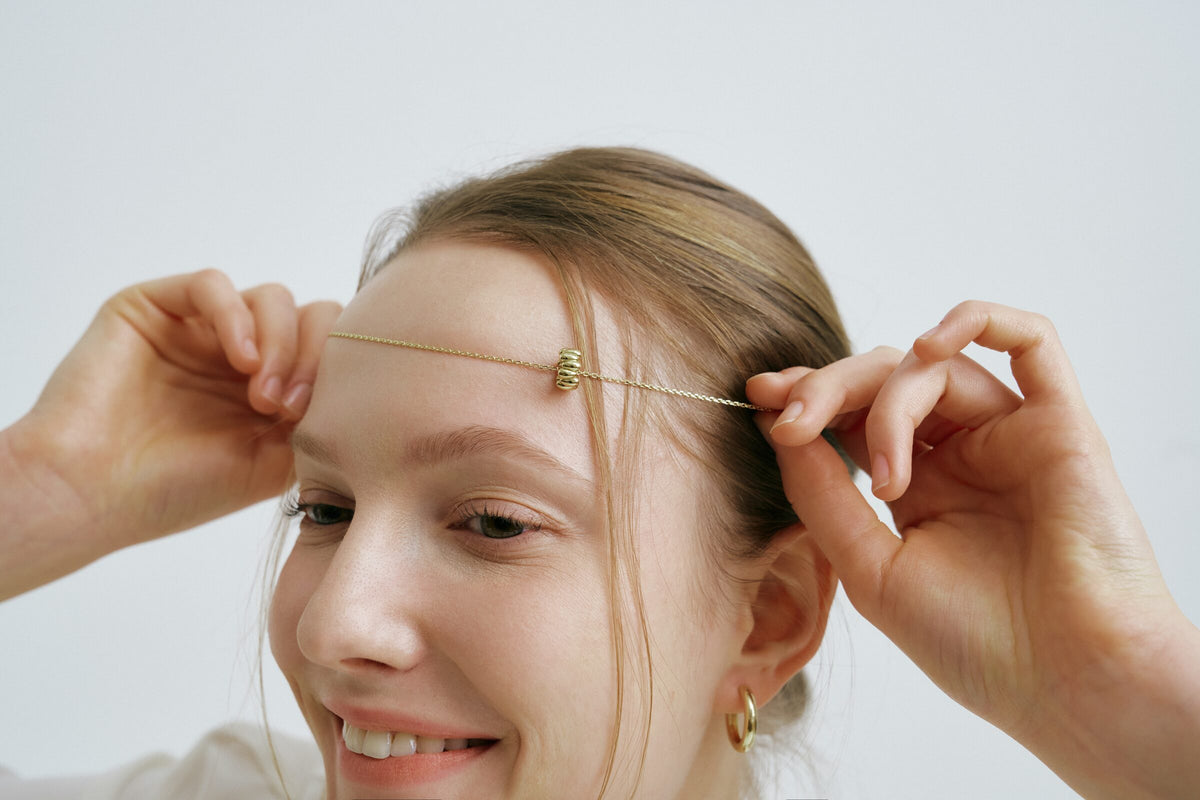 A woman smiles and holds a chain across her forehead with both hands, a ridged charm at the centre and a hoop earring.