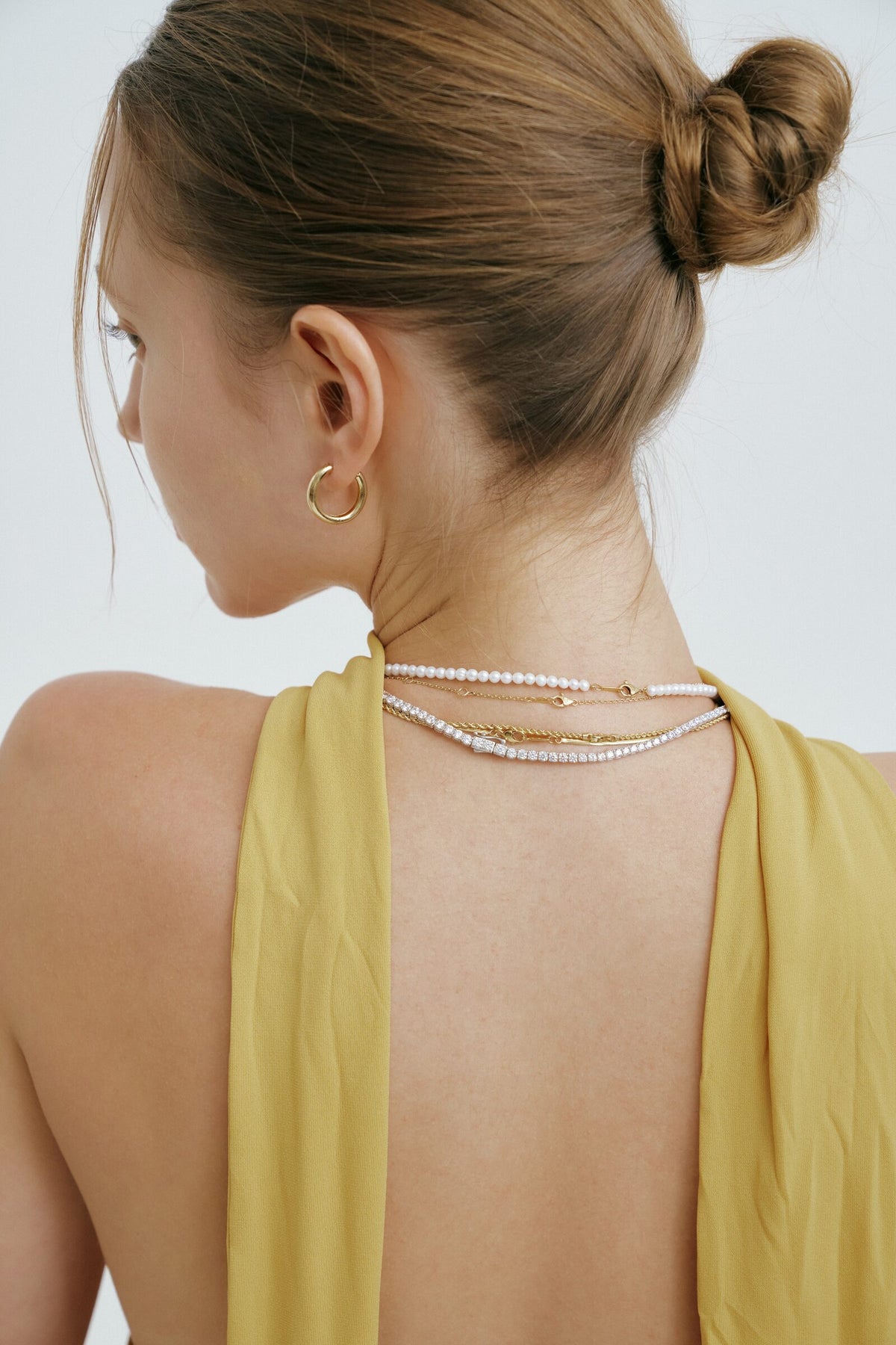 A woman seen close from behind in a backless top, pearl strands, a line of clear stones and a ridged chain at her nape.