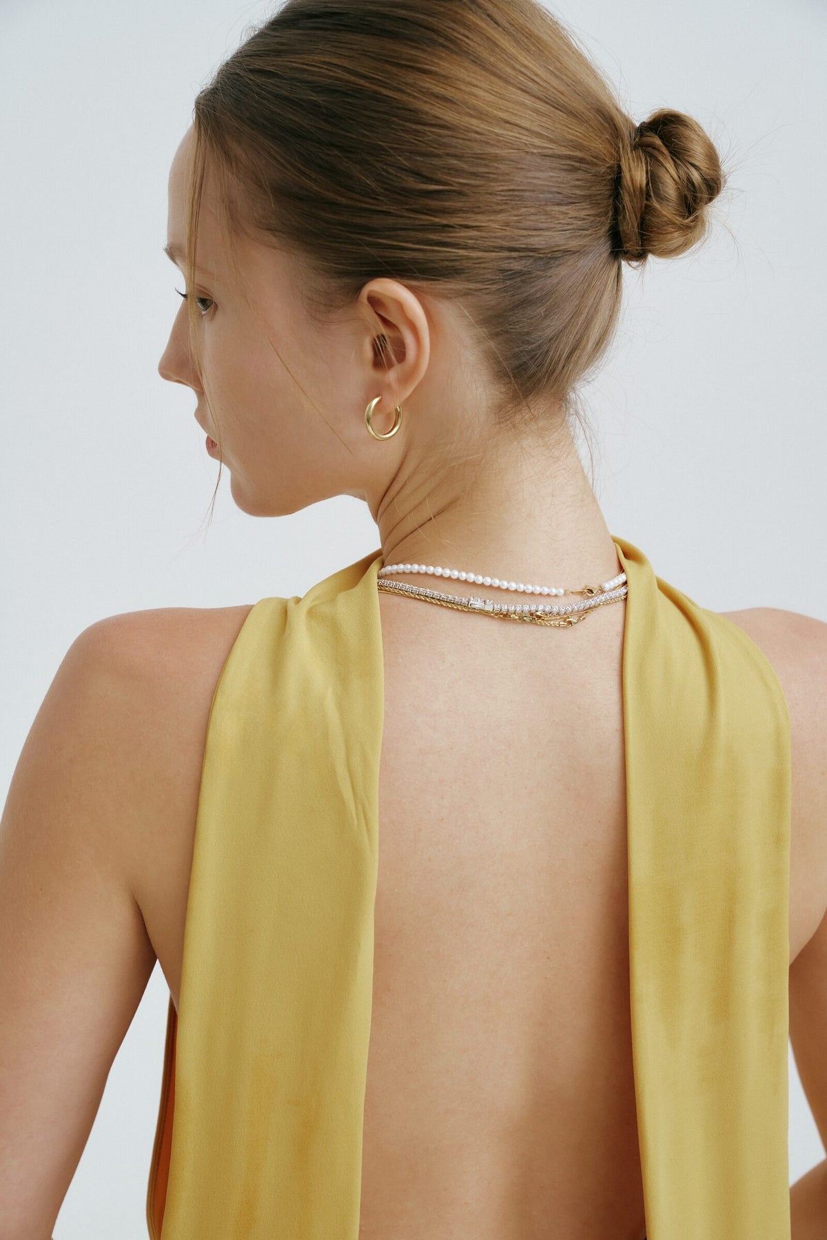 A woman seen from behind, hair in a bun, a pearl strand, a line of clear stones and two chains at her nape, a hoop earring.