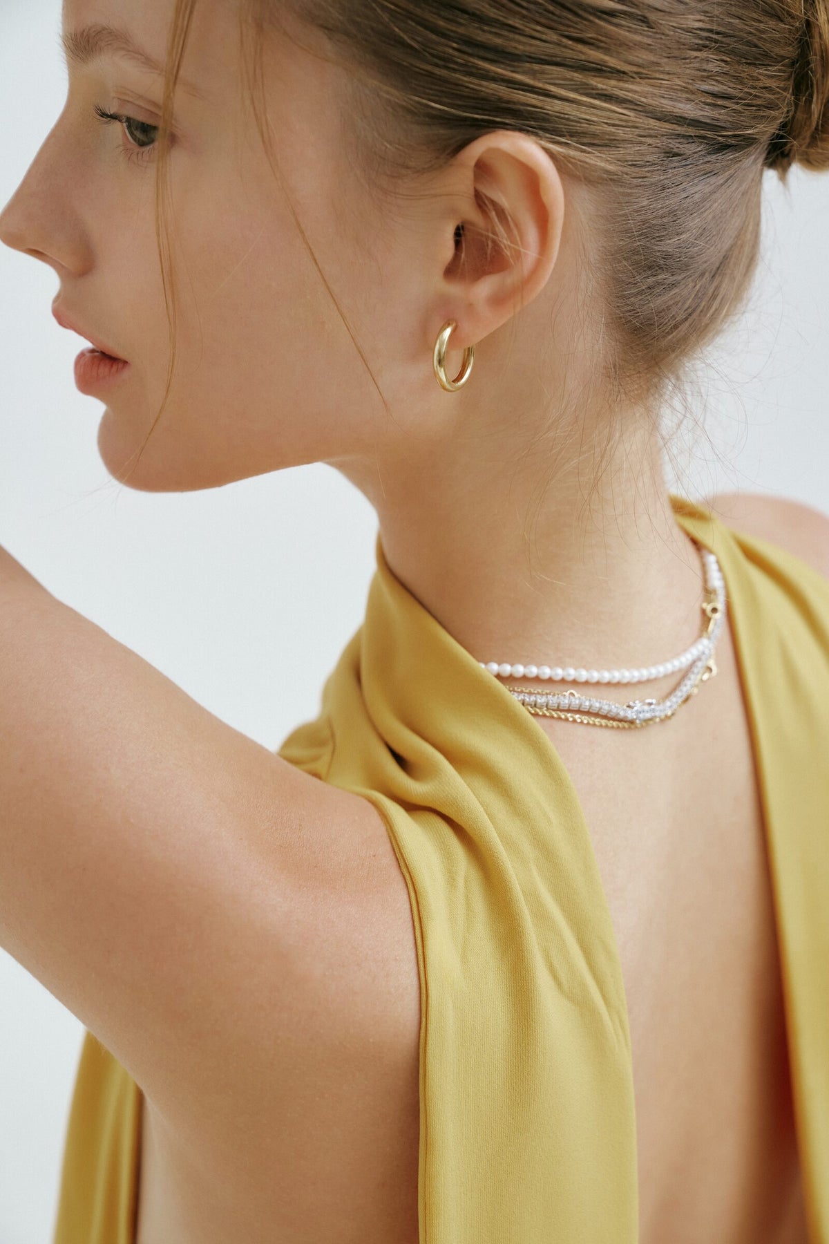 In profile over her shoulder, thick gold hoops and the necklace clasp at her neck.