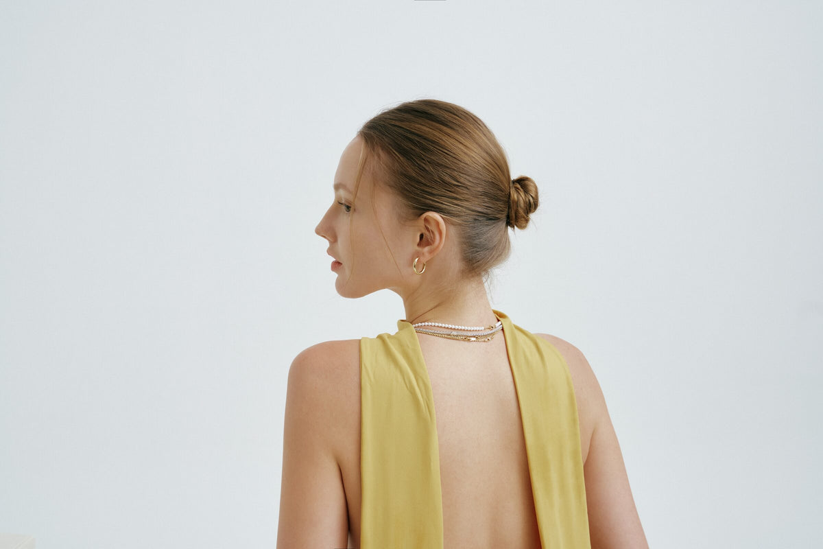 A woman in a backless top, seen in profile from a distance, hair in a bun, pearl strands at her nape and a hoop earring.