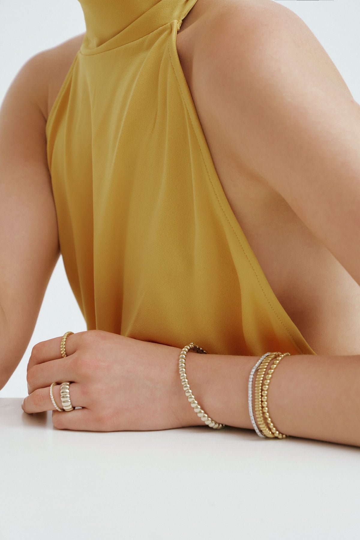 Arms on the table in a marigold halter, beaded and tennis bracelets stacked, a thin pave band and a Brioche Ring.