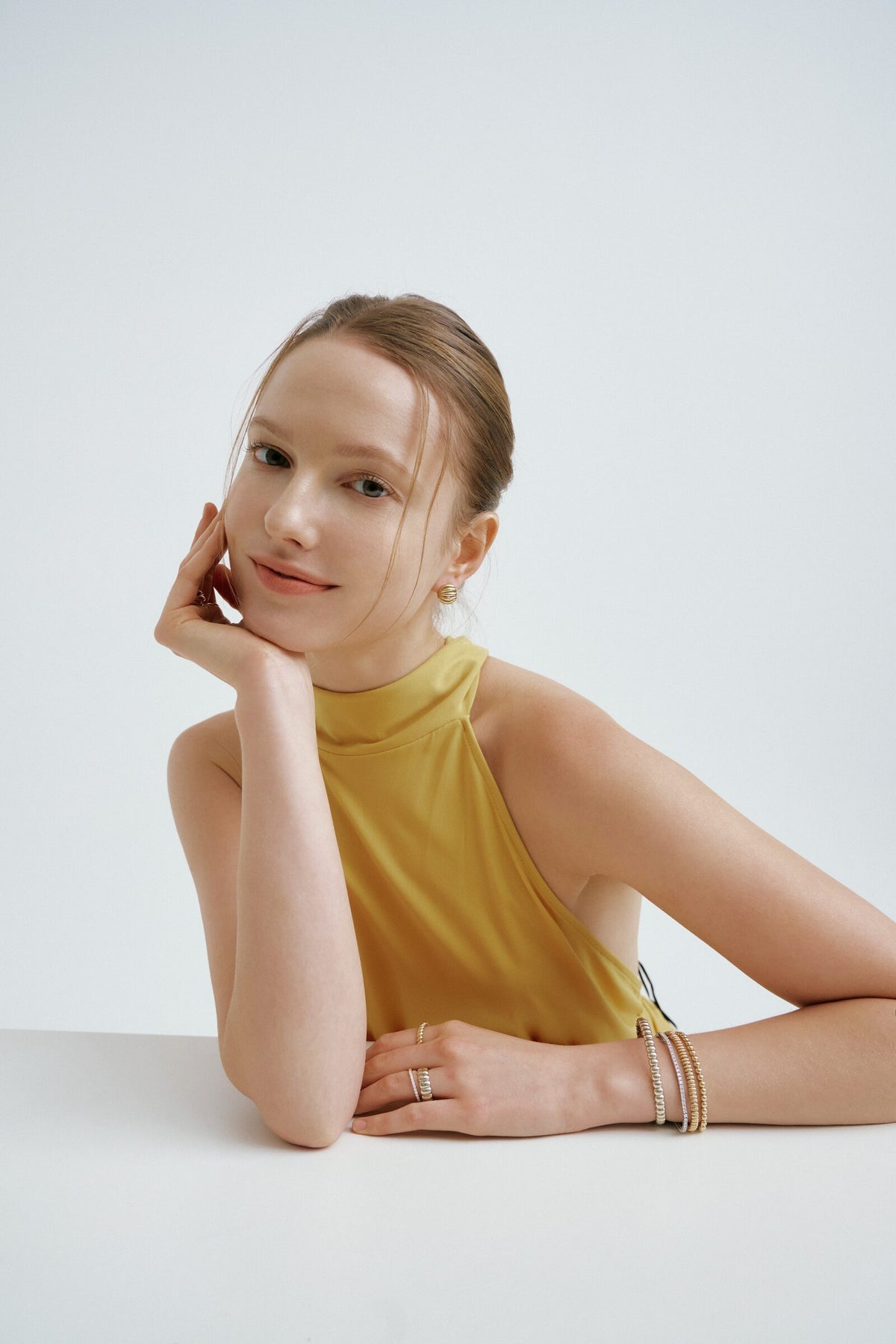 A woman leans on a table with her cheek on her hand, ridged earrings, slim rings and a stack of ridged bangles at her wrist.