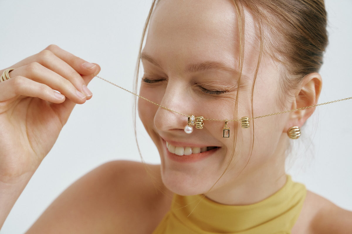 A woman laughs, eyes closed, holding a chain across her face hung with charms: a pearl, ridged beads and a small frame.