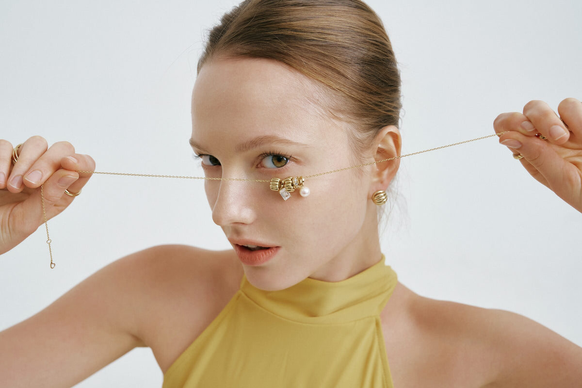 Wide frame: she pulls a charm necklace straight across her face with both hands.