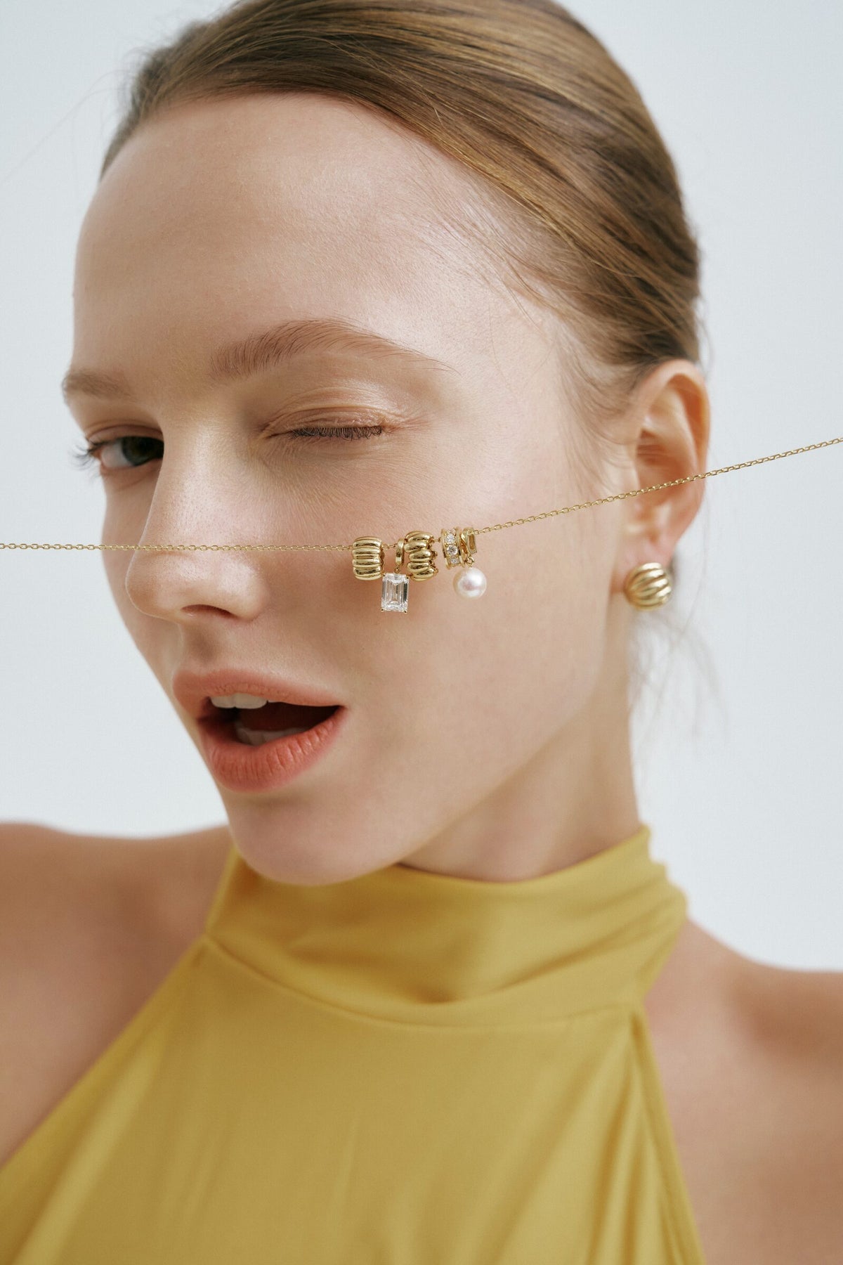 A wink, the charm chain stretched across her cheek, a ribbed gold earring