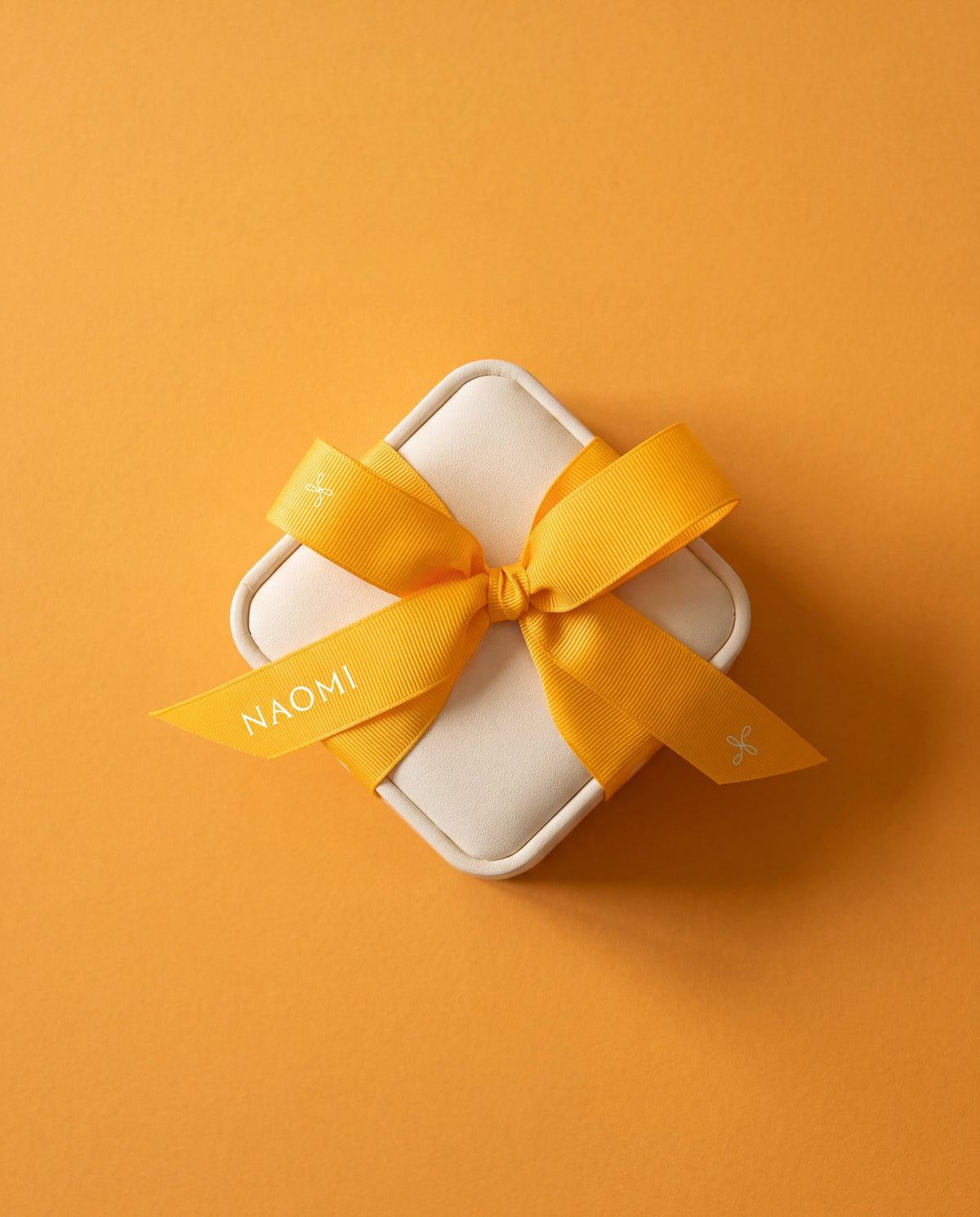 An ivory jewelry case tied with a marigold ribbon printed with the NAOMI wordmark