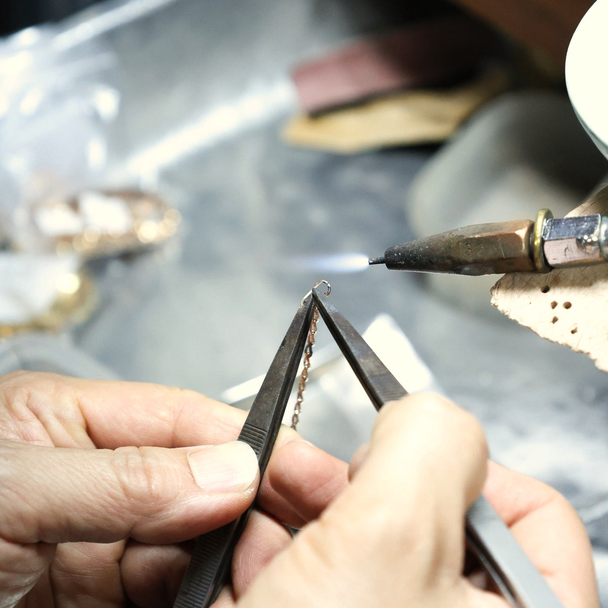 Two hands at a jeweller's bench, holding a gold chain in tweezers under a fine torch
